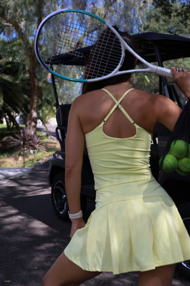 Person in a yellow skirt and tank top holding a tennis racket and balls, standing next to a golf cart.